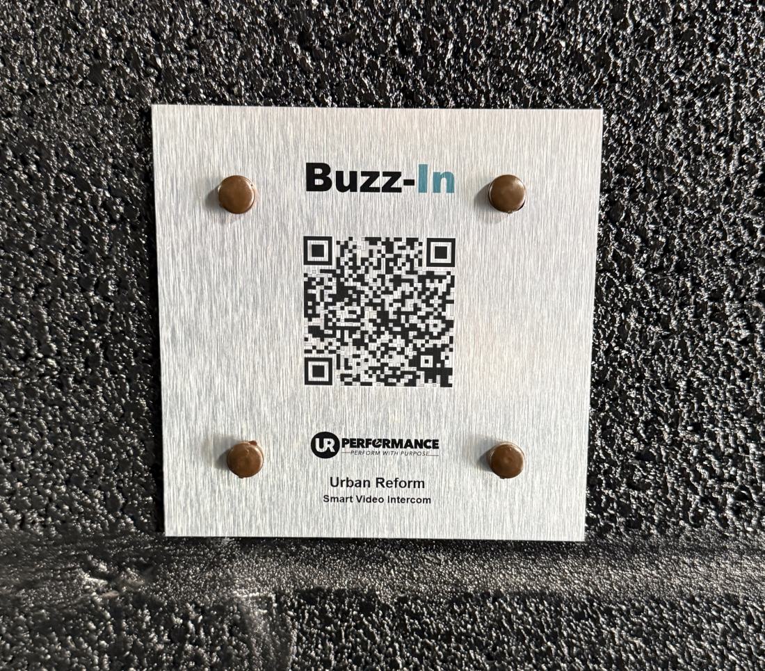 The Buzz-In QR code placard, a brushed metal plate bolted to the wall beside a communal entrance.